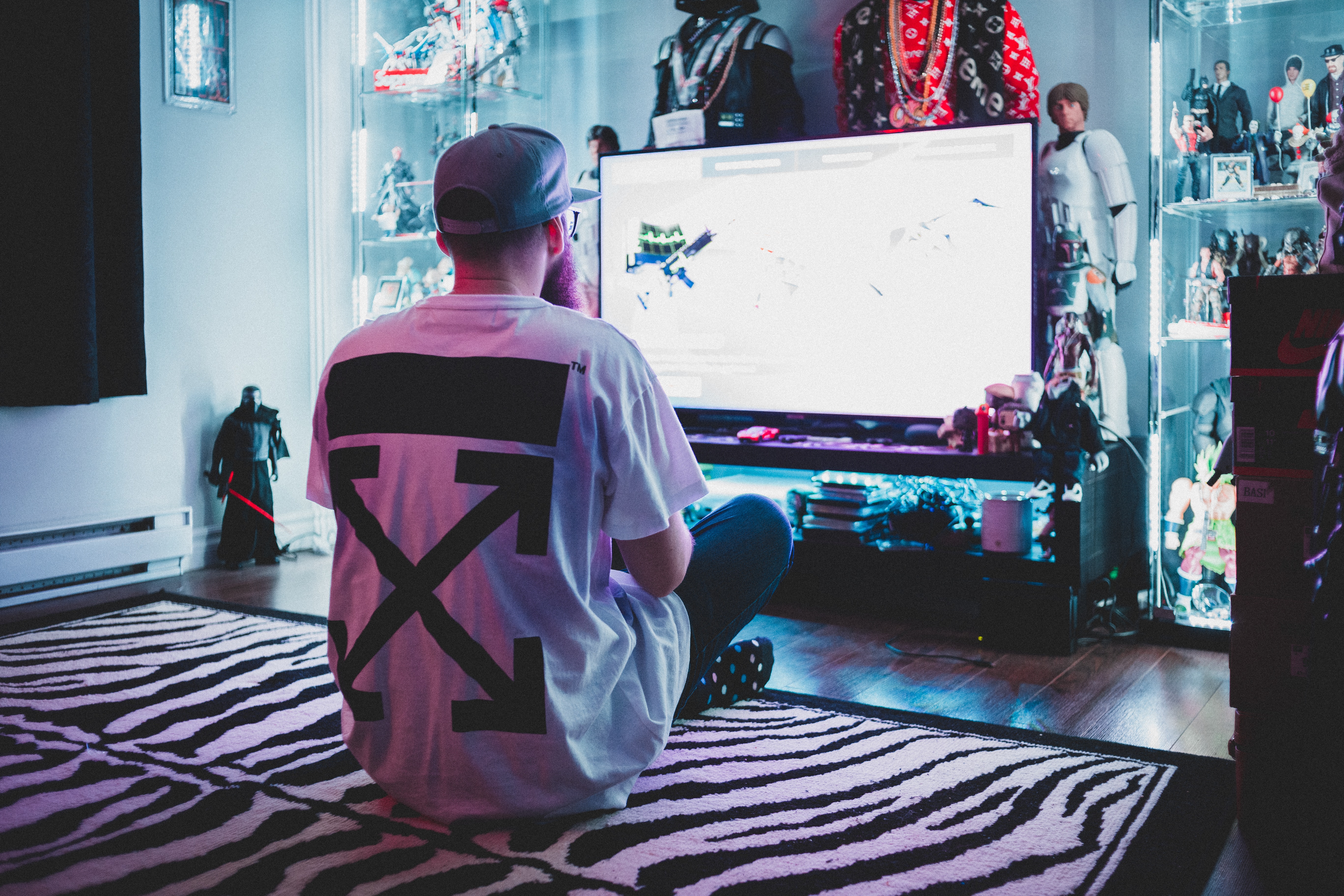 Free Back view of anonymous geek male in casual clothes and cap sitting on carpet and playing console video game in living room full of toys and stuff from comic Stock Photo