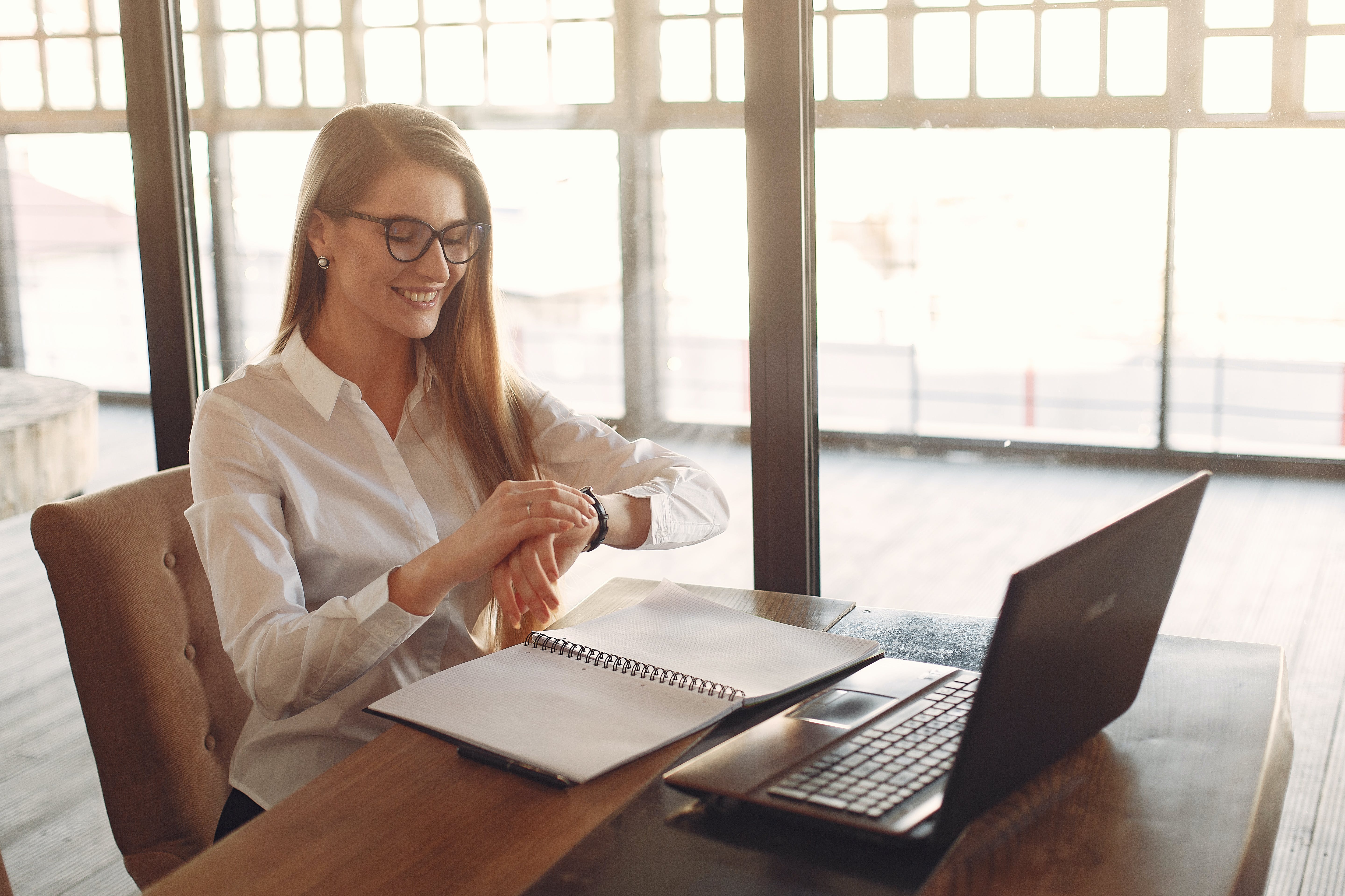 Smiling young female entrepreneur checking time on wristwatch while working on laptop in modern workspace